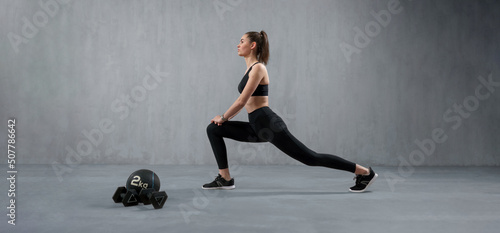 Young athletic woman in sportswear doing squat using dumbbells, posing in studio with grey background.