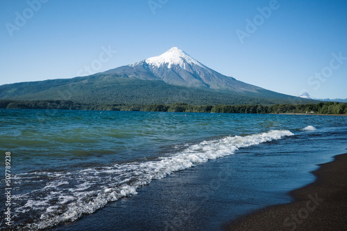 View of Osorno Volcano from shore of Lago Llanquihue Lake, Los Lagos Province, Chile. Volcano with snow-covered peak and forests on slopes, waves on lake shore and black sand beach in foreground photo