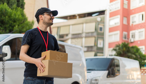 Repartidor en la ciudad con una expresión feliz, entregando un paquete, con la furgoneta de reparto al fondo.. Fotografía horizontal. © Ezequiel Martínez