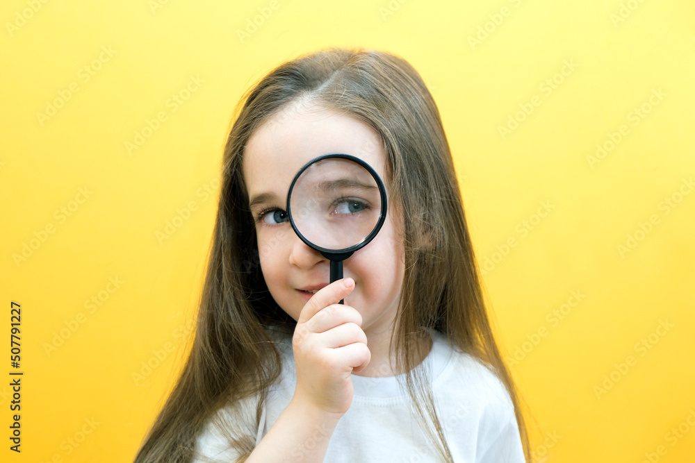Positive curious schoolgirl in casual clothes looks at the camera through a magnifying glass, on a yellow background. Positive curious schoolgirl in casual clothes looks at the camera through a magnifying glass, on a yellow background.