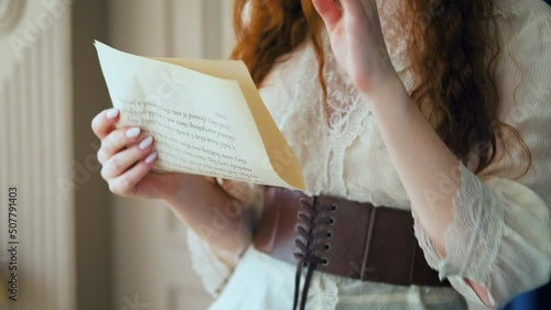 Red-haired fantasy business woman portrait close-up holds documents in hands face cropped. Medieval girl princess reads love letter. Vintage dress long red hair, brown suit white blouse, old room. 4k photo