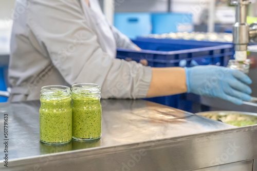 Pesto sauce Industrial process. High quality sauce production. Worker operator at the food factory behind the machine pours pesto sauce into jars. Italian basil taste in Pesto sauce