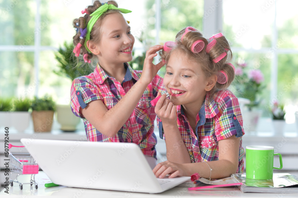 Cute twin girls in hair curlers with laptop at home appying lipstick Cute twin girls in hair curlers with laptop at home appying lipstick