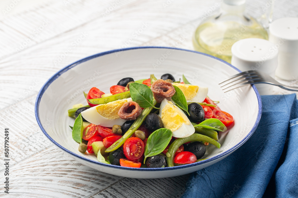 Close-up of french Nicoise salad with anchovy, green beans, tomatoes, eggs, black olives, capers on white plate and white wooden background. Healthy food, no carb diet concept.