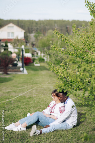 Interracial couple sits on grass in spring garden dressed in Ukrainian embroidered shirts.