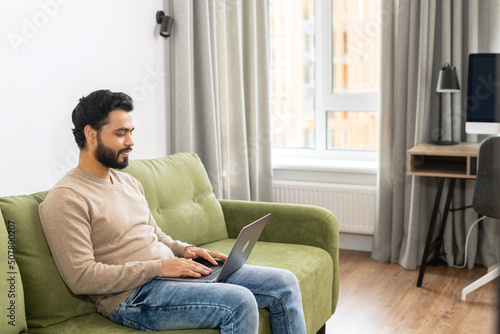 Positive Hindi man in smart casual clothes using computer while sitting at the sofa in his flat. Young Indian male student watching webinars, educational courses, learning on the distance