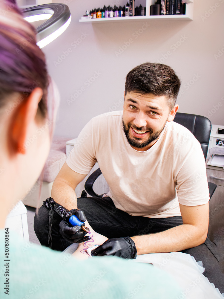 Work as a tattoo artist in a tattoo parlor. a tattoo artist in black gloves is working on a tattoo on the leg of a young woman and looking at the client and smiling Work as a tattoo artist in a tattoo parlor. a tattoo artist in black gloves is working on a tattoo on the leg of a young woman and looking at the client and smiling