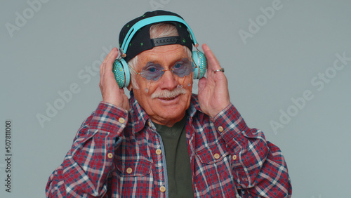 Portrait of senior man 80s years old in checked shirt listening music via headphones and dancing disco fooling around having fun expressive gesticulating hands. Old grandfather on gray wall background