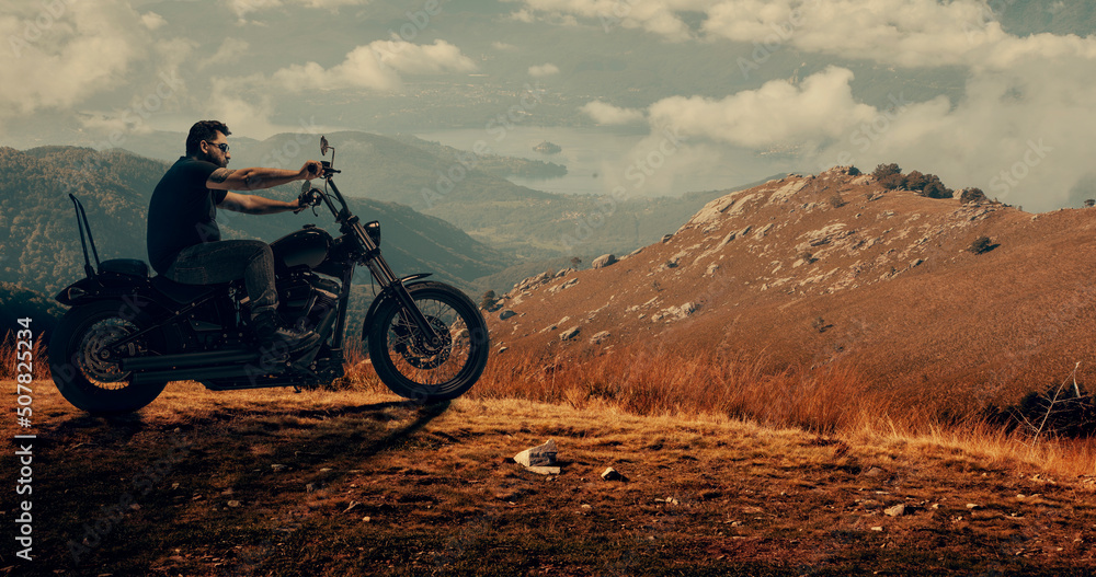 Handsome muscular man, professional biker on cool bike has a rest at mountain road at summer sunset. Concept of dream, travel, style, techno and achievements Handsome muscular man, professional biker on cool bike has a rest at mountain road at summer sunset. Concept of dream, travel, style, techno and achievements