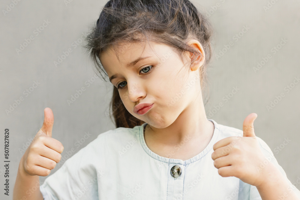 A little girl emotionally pouted her lips and shows a thumbs up on a gray background, childish behavior and imitation of parents A little girl emotionally pouted her lips and shows a thumbs up on a gray background, childish behavior and imitation of parents