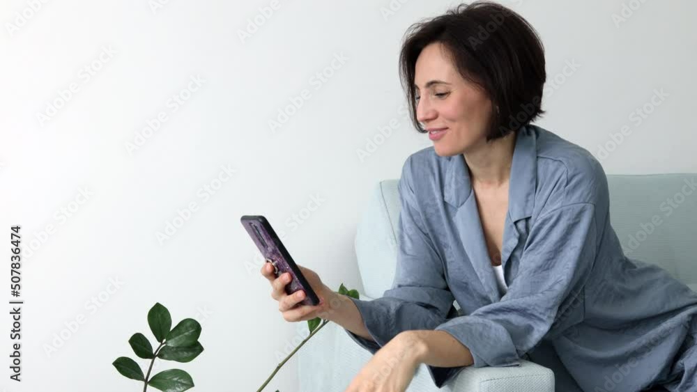 Smiling european female sitting  on sofa hold smartphone talk by video call wave hand to camera. Positive young mixed race woman contact call friend by video link chat using device webcam