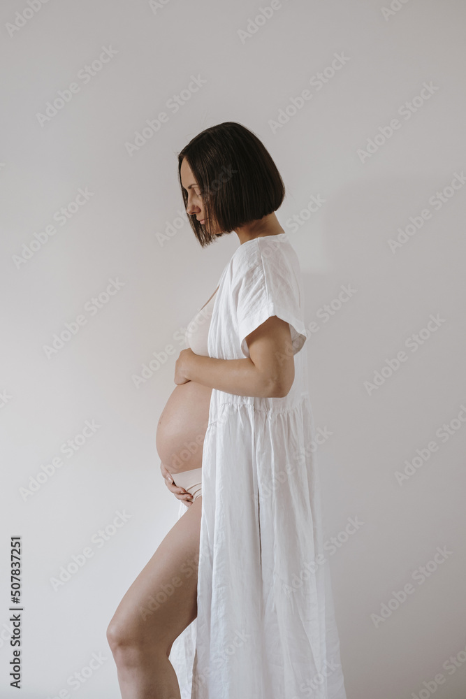 Happy beautiful young pregnant woman in white robe stays against white wall Happy beautiful young pregnant woman in white robe stays against white wall
