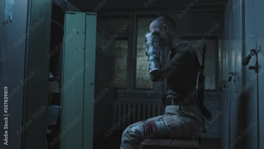 Medium long side view of injured female soldier with shaved head sitting in abandoned dark locker room, patching up wounds with duct tape and available means Medium long side view of injured female soldier with shaved head sitting in abandoned dark locker room, patching up wounds with duct tape and available means