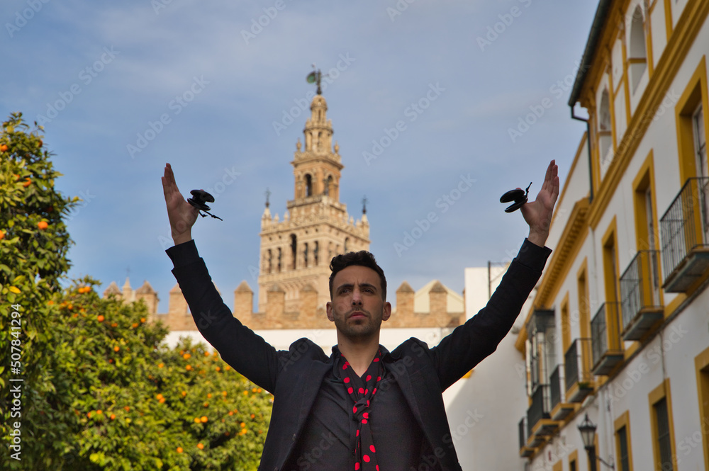 Fototapeta premium gypsy man dancing flamenco, dressed in black with castanets is posing with his arms up among orange trees and in the background the giralda of seville. Flamenco cultural heritage of humanity