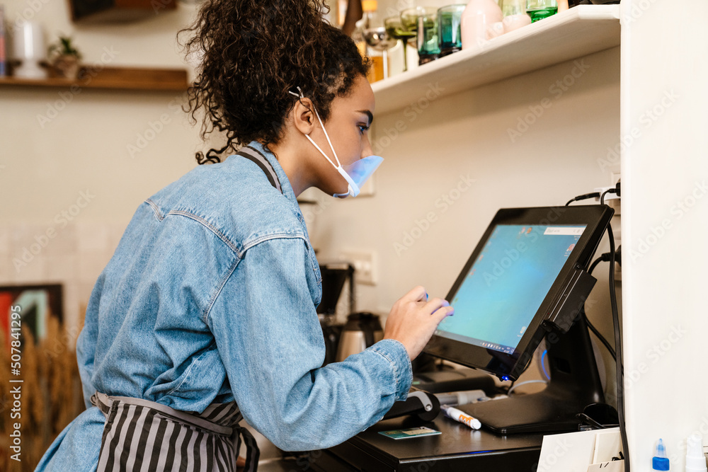 Black barista woman wearing face mask working with cash register in cafe