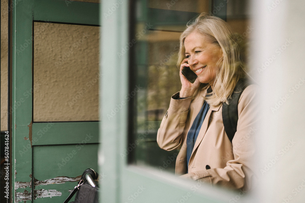 Smiling businesswoman talking on smart phone seen through glass window Smiling businesswoman talking on smart phone seen through glass window