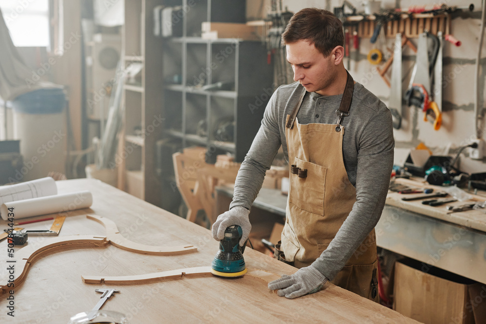 Portrait of young carpenter sanding piece of handcrafted wooden furniture in hazy workshop, copy space