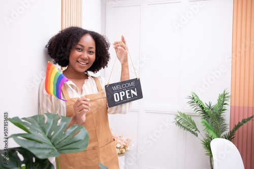 Beautiful African American woman business owner holding a store opening sign and holding a rainbow flag supporting LGBTQ+ gender equality bags homosexuality, success, confidence.
