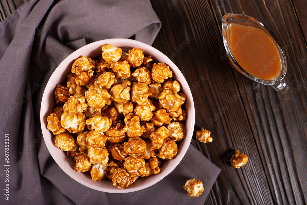 Clay bowl with caramel popcorn on wooden background flat lay