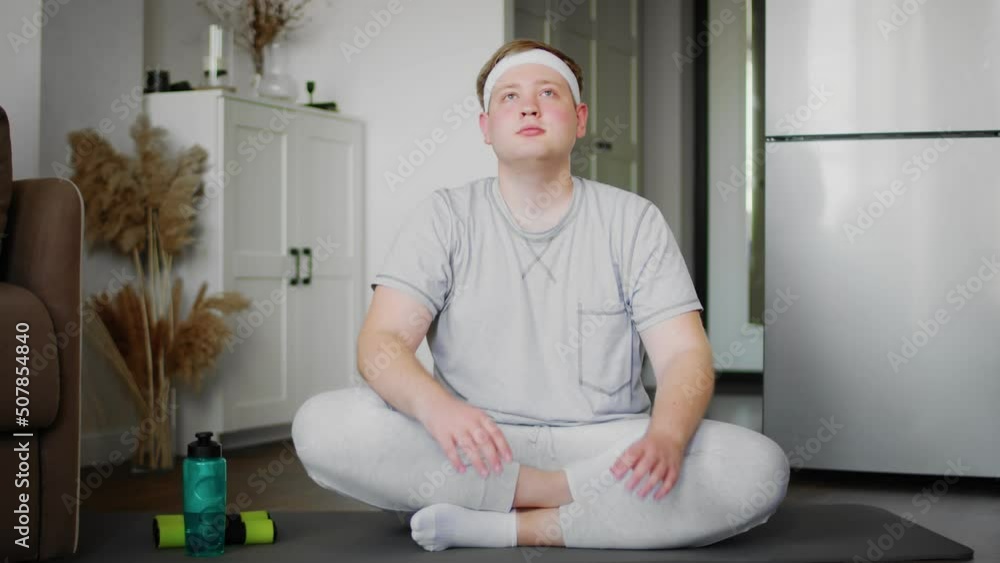 The guy stretches his neck while sitting on the floor in the lotus position at home in the living room in sportswear. Sport The guy stretches his neck while sitting on the floor in the lotus position at home in the living room in sportswear. Sport