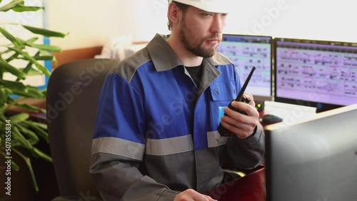 Serious Caucasian unshaven worker in overalls and helmet sitting in the control room gives instructions using a radio station. Automated workplace of a dispatcher or operator of a modern enterprise photo