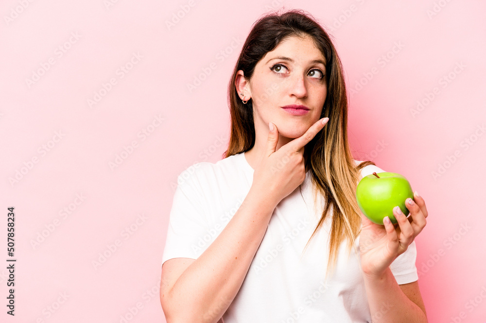 Young caucasian woman holding an apple isolated on pink background looking sideways with doubtful and skeptical expression. Young caucasian woman holding an apple isolated on pink background looking sideways with doubtful and skeptical expression.