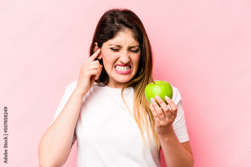 Young caucasian woman holding an apple isolated on pink background covering ears with hands. Young caucasian woman holding an apple isolated on pink background covering ears with hands.