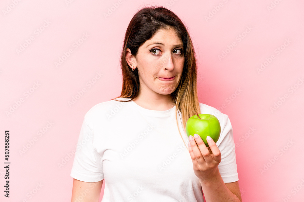 Young caucasian woman holding an apple isolated on pink background confused, feels doubtful and unsure. Young caucasian woman holding an apple isolated on pink background confused, feels doubtful and unsure.