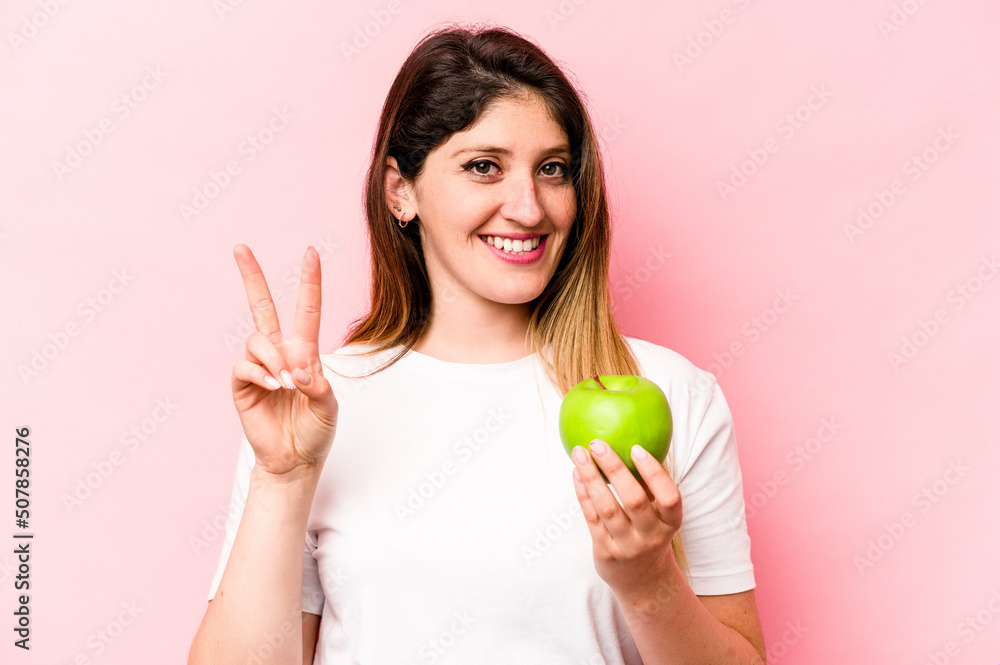 Young caucasian woman holding an apple isolated on pink background showing number two with fingers. Young caucasian woman holding an apple isolated on pink background showing number two with fingers.