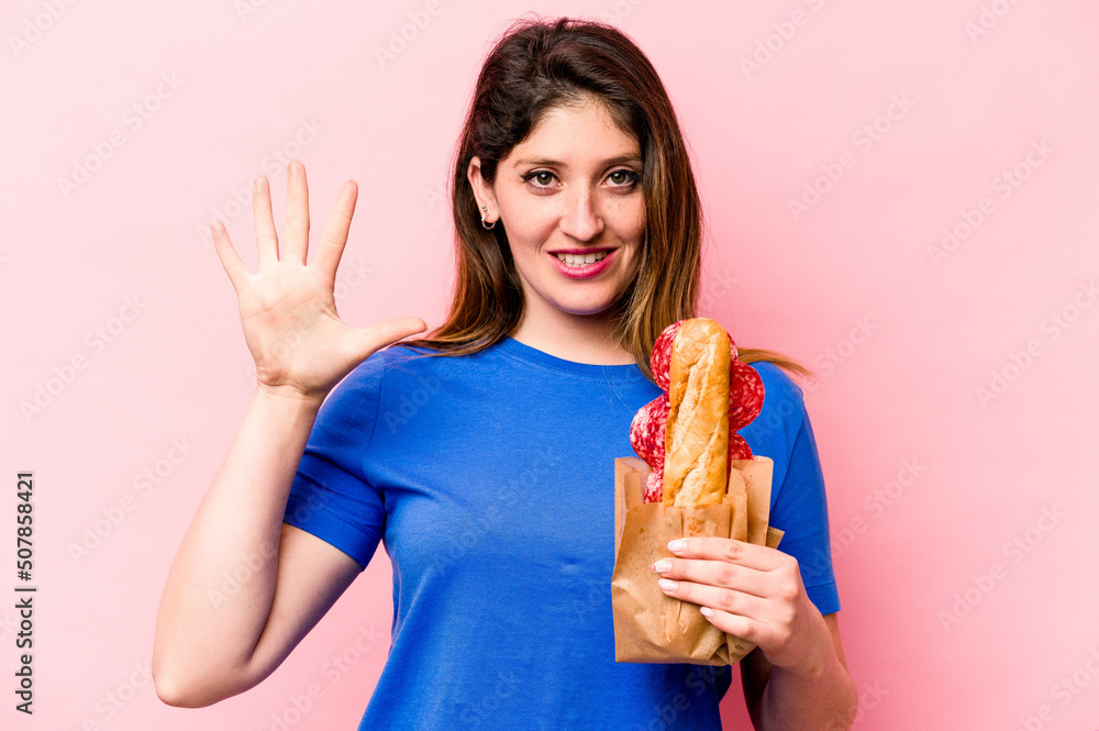 Young caucasian woman eating a sandwich isolated on pink background smiling cheerful showing number five with fingers.