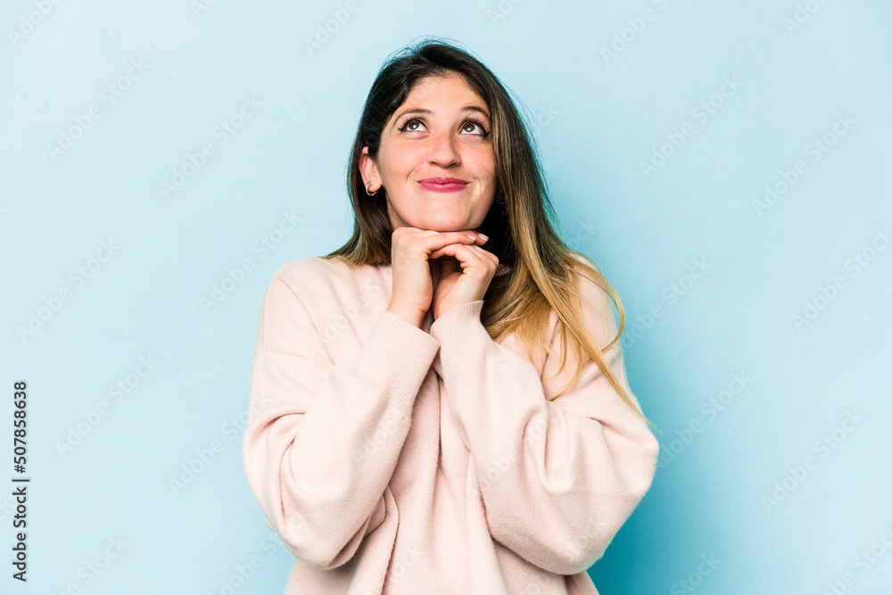 Young caucasian woman isolated on blue background keeps hands under chin, is looking happily aside. Young caucasian woman isolated on blue background keeps hands under chin, is looking happily aside.
