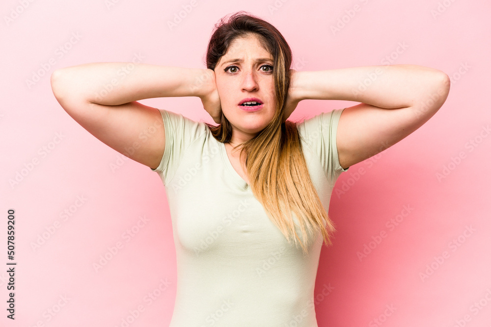 Young caucasian woman isolated on pink background covering ears with hands trying not to hear too loud sound. Young caucasian woman isolated on pink background covering ears with hands trying not to hear too loud sound.