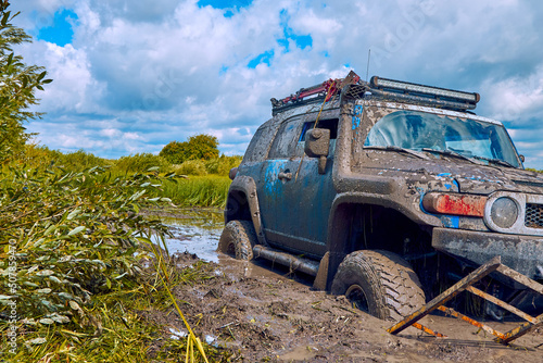 Blue 4x4 car or 4WD car with wheels in mud on the off-road on a background of greenery and blue sky with clouds. An off-road car is stuck in a puddle of mud and is trying to leave using a ladder. © Aleksandr
