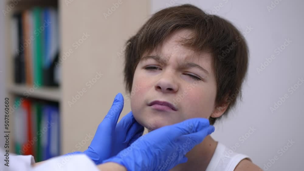 Headshot portrait of sad Caucasian boy with painful throat sitting in hospital as pediatrician doing glands palpation in slow motion. Upset little patient undergoing medical checkup indoors Headshot portrait of sad Caucasian boy with painful throat sitting in hospital as pediatrician doing glands palpation in slow motion. Upset little patient undergoing medical checkup indoors