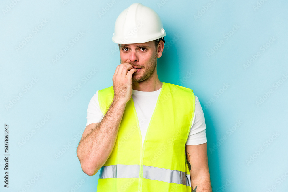 Young laborer caucasian man isolated on blue background biting fingernails, nervous and very anxious.