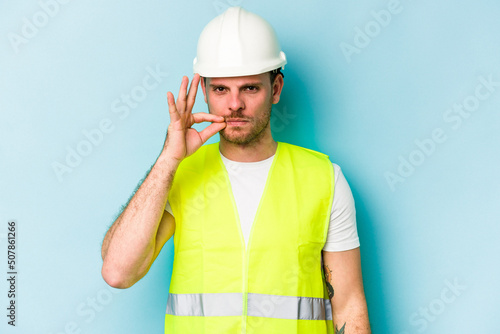 Young laborer caucasian man isolated on blue background with fingers on lips keeping a secret.