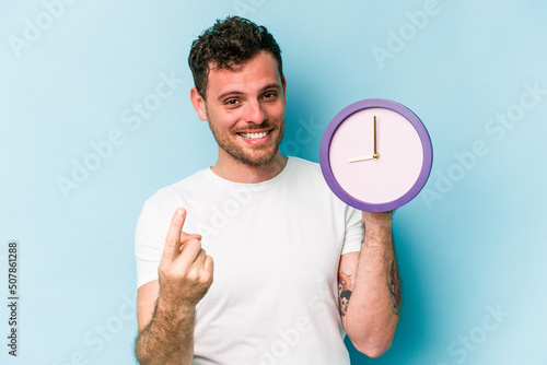 Young caucasian man holding a clock isolated on blue background pointing with finger at you as if inviting come closer.