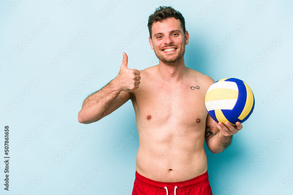 Young caucasian man playing volley isolated on blue background smiling and raising thumb up