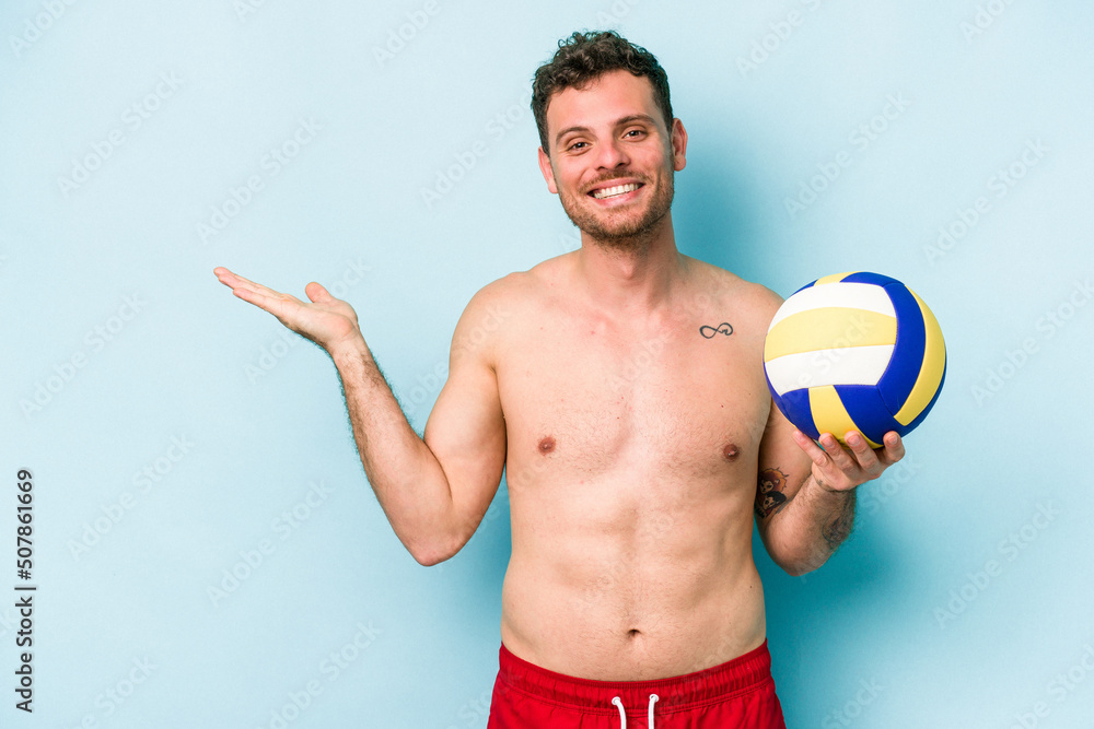Young caucasian man playing volley isolated on blue background showing a copy space on a palm and holding another hand on waist.