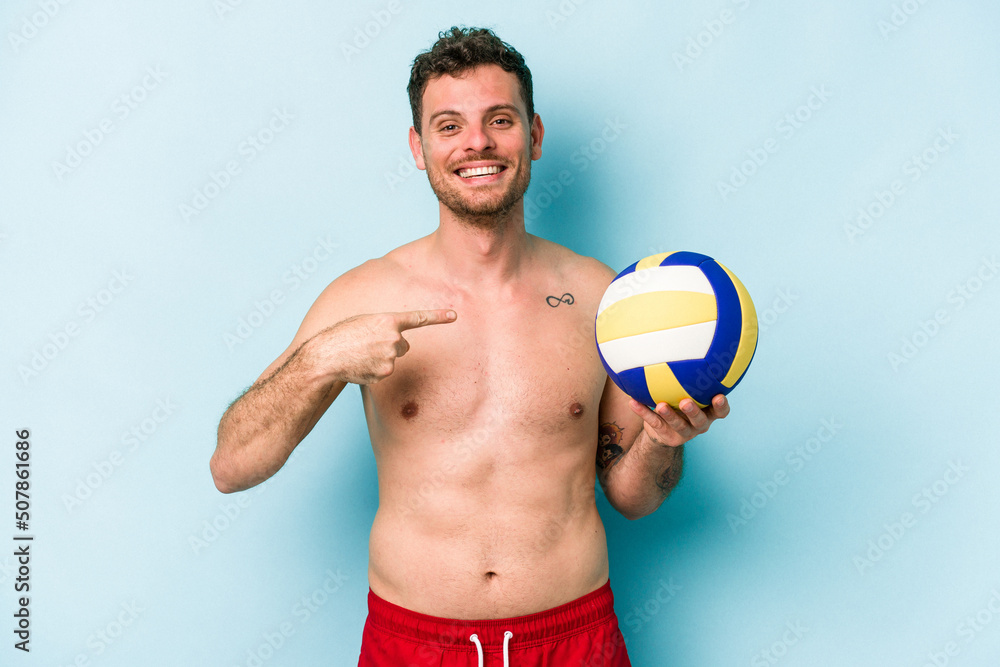 Young caucasian man playing volley isolated on blue background person pointing by hand to a shirt copy space, proud and confident