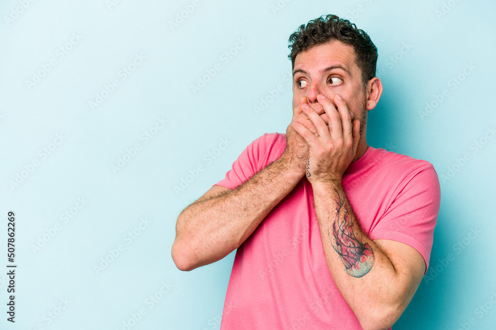 Young caucasian man isolated on blue background thoughtful looking to a copy space covering mouth with hand. Young caucasian man isolated on blue background thoughtful looking to a copy space covering mouth with hand.