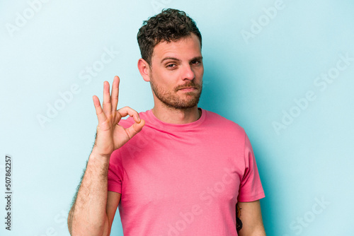 Young caucasian man isolated on blue background winks an eye and holds an okay gesture with hand.