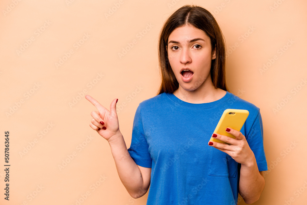Young caucasian woman holding mobile phone isolated on beige background pointing to the side Young caucasian woman holding mobile phone isolated on beige background pointing to the side