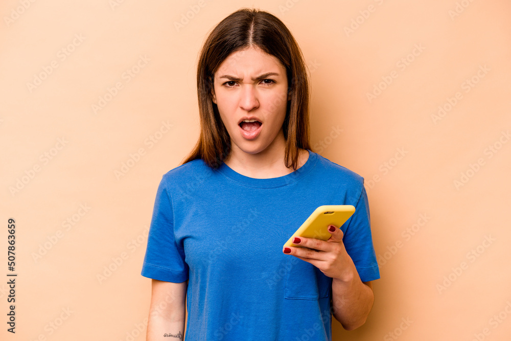 Young caucasian woman holding mobile phone isolated on beige background screaming very angry and aggressive. Young caucasian woman holding mobile phone isolated on beige background screaming very angry and aggressive.