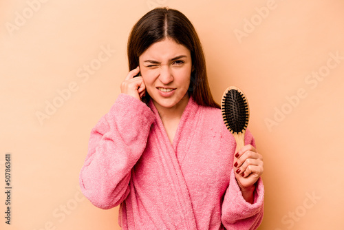 Young hispanic woman holding hairbrush isolated on beige background covering ears with hands.