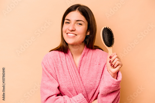 Young hispanic woman holding hairbrush isolated on beige background laughing and having fun.