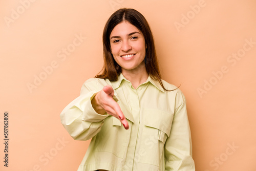 Young hispanic woman isolated on beige background stretching hand at camera in greeting gesture.