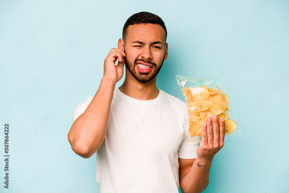 Young hispanic man holding a bag of chips isolated on blue background covering ears with hands. Young hispanic man holding a bag of chips isolated on blue background covering ears with hands.
