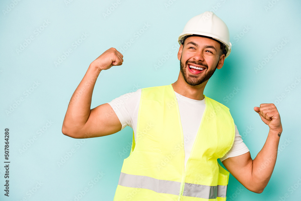 Young laborer hispanic man isolated on blue background raising fist after a victory, winner concept.