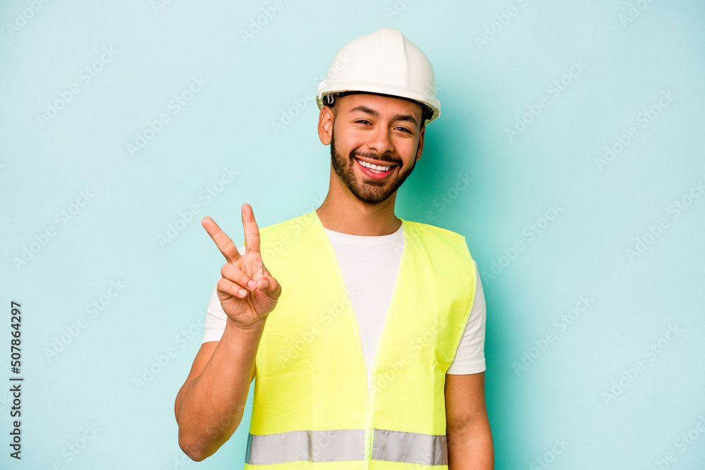 Young laborer hispanic man isolated on blue background joyful and carefree showing a peace symbol with fingers.